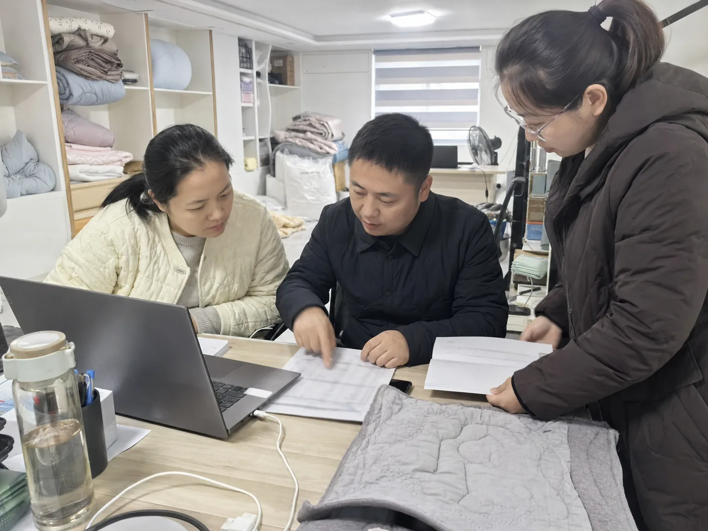 三名家纺业务人员在办公桌旁核对绗缝样品、电脑页面和工艺资料